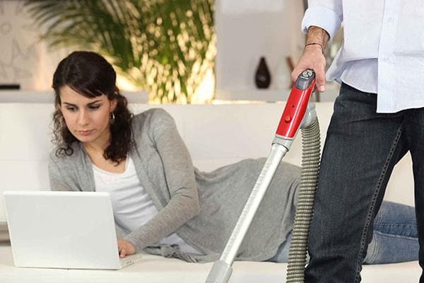 Frau liegt mit Laptop auf dem Sofa, während der Mann staubsaugt